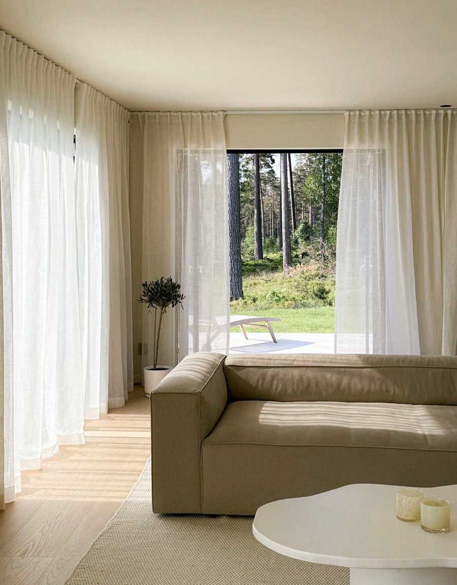 Modernt vardagsrum med beige soffa, vitt soffbord och stora fönster med ljusbeiga gardiner